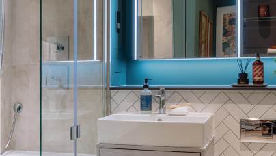 Modern bathroom with a glass shower, white sink with a blue soap dispenser, and a large mirror. The walls are painted teal and tiled with a chevron pattern.