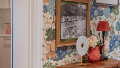 Framed black and white photo on a wallpapered wall with nature and animal patterns, beside a wooden table with flowers, a lamp, and a decorative object. Kitchen visible through an open door.