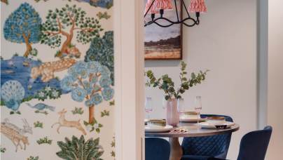 A dining area with a round table set for a meal, four blue chairs, and a vase with leaves. A wall with a colorful nature-themed wallpaper is partially visible.