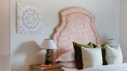 Bedroom with a decorative pink patterned headboard, two green pillows, and a wooden bedside table with a lamp. Wall art with an intricate white pattern hangs above the bed.