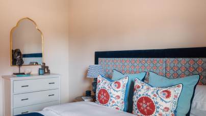 A bedroom with a bed featuring patterned pillows and a blue headboard. A white dresser is topped with a decorative mirror and small items. The room has a soft, neutral color scheme.