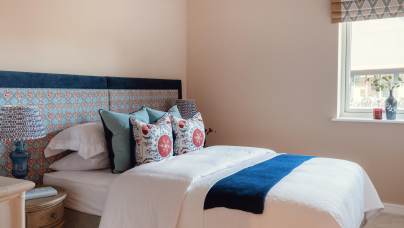 A neatly made bed with patterned pillows and a blue throw, next to a wooden side table with a lamp, in a room with beige walls and a window with a Roman shade.