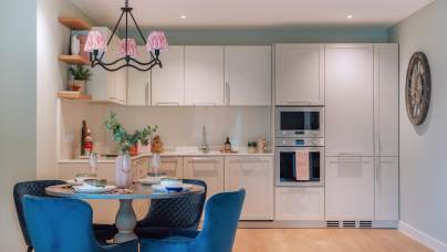 Modern kitchen featuring white cabinets, built-in ovens, a round dining table with blue chairs, and a black chandelier with pink shades. A large wall clock is visible.