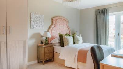 A neatly arranged bedroom featuring a bed with orange patterned headboard, green and white pillows, bedside table with lamps, and a window with drapes. A decorative wall piece hangs above.