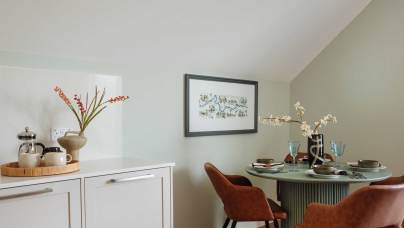 A cozy dining area with a round table set for four, two brown chairs, and minimalist decor. Nearby, a countertop holds a wooden tray with a French press and mugs. Neutral tones dominate the space.