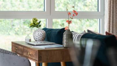 A cozy living room corner with a wooden table, two vases with flowers, a glass, and a laptop. A green couch sits against large windows showing a blurred outdoor view.