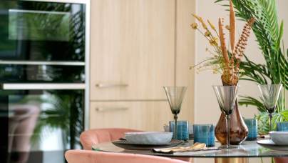 Dining table set with dishes and glasses, featuring a vase with dried flowers. Pink chairs surround the table. Wooden cabinets and green plants are in the background.