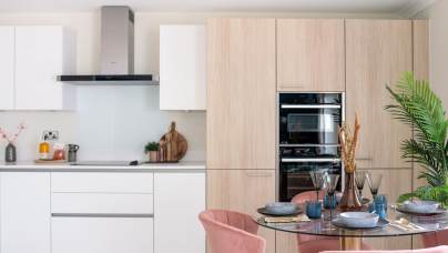 Modern kitchen with white cabinets, light wood paneling, and a stainless steel oven. There is a round dining table with pink chairs and tableware. A large green plant is in the corner.
