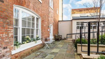 A small courtyard with stone paving, a white window, two metal chairs, a table, and a black metal gate.