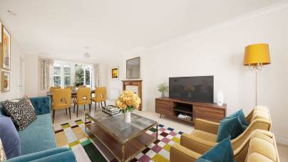 Spacious living room with a blue sofa, yellow armchairs, a large TV, and a dining table with yellow chairs. A colorful rug covers the floor, and a bouquet of flowers is on the glass coffee table.