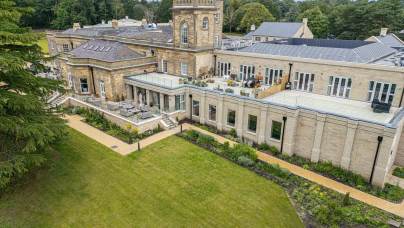 Aerial view of a large, historic stone building with a central tower and garden. The structure has multiple windows and adjacent walkways, surrounded by lush green lawns and trees.