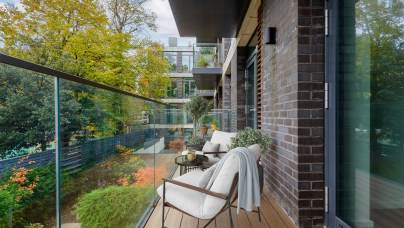 Modern balcony with glass railing, two chairs, a table, and potted plants. Trees with autumn foliage visible in the background.