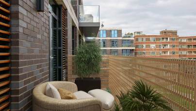 Balcony view with wicker seating, potted plants, and nearby apartment buildings under a cloudy sky.