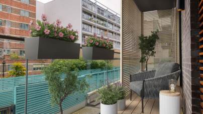 Modern balcony with two planters of pink flowers, a gray cushioned chair, a small round table with a candle, and a potted plant. Wood slat wall and glass railing overlook neighboring buildings.