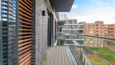 Balcony with wooden flooring and glass railing, flanked by a brick building with wooden slats on the left. Overlooks modern apartment buildings and a green rooftop under a blue sky.