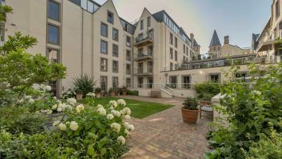 A courtyard of an apartment complex with a brick pathway, benches, green lawn, and white flowering plants.