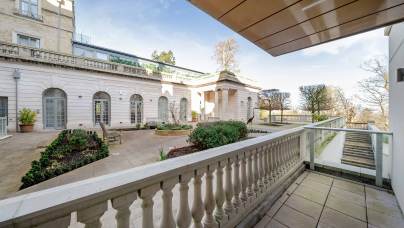 View of a spacious courtyard featuring a stone balustrade, surrounded by a classical building facade with arched doors and a neatly arranged garden.