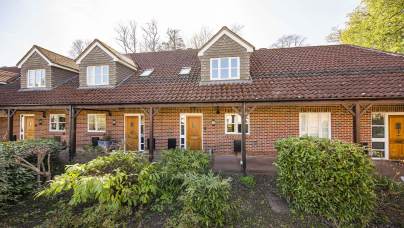 A row of brick terraced houses with tiled roofs, front porches, and wooden doors, bordered by green shrubs and bushes.