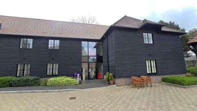 A two-story building with dark wooden siding, a glass entryway, brick trim, benches, and landscaped greenery in front.