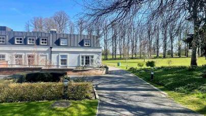 A paved driveway curves past a white house with a black roof, surrounded by manicured lawns, bushes, and tall leafless trees under a clear blue sky.