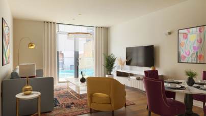 A modern living room features a gray sofa, yellow and pink chairs, a flat-screen TV, and colorful wall art. Large windows overlook a balcony with a pool view.
