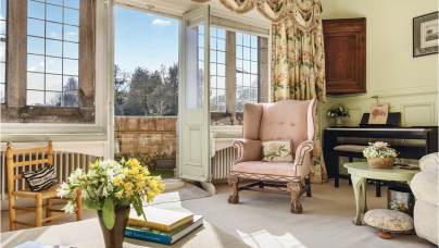 A bright, vintage-style living room with floral upholstery and a view of a garden through open French doors. A pink armchair sits next to a piano, with books and flowers on a table.