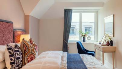 A neatly arranged bedroom with a bed, colorful pillows, a window with curtains, a small desk with flowers, and a blue chair.