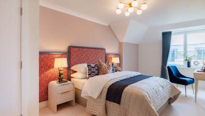 A bedroom featuring a neatly made bed with red patterned headboard, two bedside tables with lamps, a blue chair, a desk near a window, and a modern ceiling light.