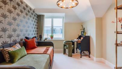 Room with a daybed, decorative pillows, a desk, chair, and lamp. Wallpaper with a leaf pattern on one wall, window with a view, and a mix of green and orange accents.