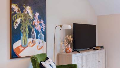 A living room with a green armchair, large floral painting, wall-mounted lamp, flat-screen TV on a white cabinet, and a decorative vase with flowers.