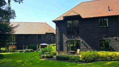 Two dark wooden buildings with red-tiled roofs sit in a landscaped garden with green grass, shrubs, and outdoor furniture on a sunny day.