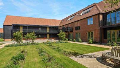 A modern brick building with balconies faces a landscaped courtyard with green lawns, young trees, and a circular wooden bench under a blue sky.