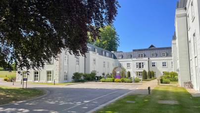 White multi-story building with black roof, surrounded by green lawn, trees, and a driveway under a clear blue sky.