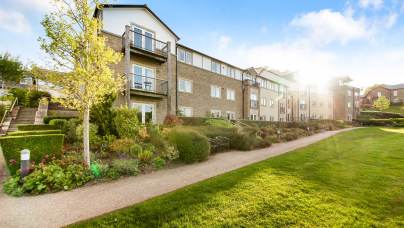 A modern, multi-story residential building with balconies and landscaped gardens, photographed on a sunny day with a clear blue sky.