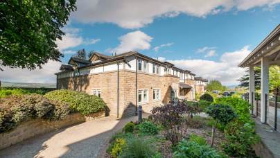 Two-story stone building with white trim, surrounded by landscaped gardens, under a partly cloudy blue sky. A paved pathway leads through the greenery.