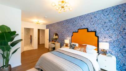 A modern bedroom with a blue patterned accent wall, orange headboard, double bed, bedside tables, a potted plant, and a small vanity area near an open doorway.