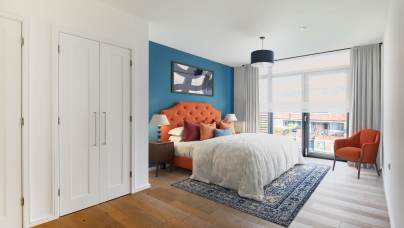 Modern bedroom with an orange upholstered headboard, blue accent wall, white bedding, patterned rug, armchair, two nightstands, and large window with sheer curtains.