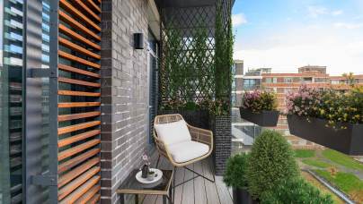 A modern balcony with a chair, small table, potted plants, and a privacy screen, overlooking a cityscape with buildings and greenery.