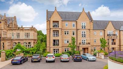 Parking lot with six cars in front of a modern stone apartment building, with an older stone building to the left and greenery around.