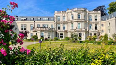 A large white historic building with ornate architecture, surrounded by a manicured garden and blooming flowers under a clear blue sky.