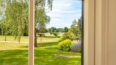 View through a window showing a well-maintained garden with green lawn, flowering shrubs, trees, and a park-like landscape under a blue sky.