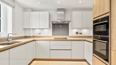 Modern kitchen with white cabinets, light wood accents, built-in double oven, electric cooktop, stainless steel hood, and a window above a single-basin sink.