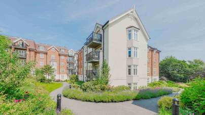A modern apartment building with balconies, surrounded by landscaped gardens and walking paths under a clear blue sky.