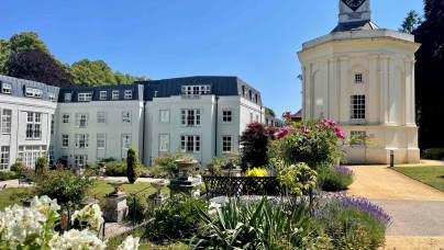 A white building with multiple windows stands beside a white clock tower, surrounded by landscaped gardens and flowering plants under a clear blue sky.