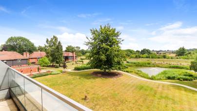 View from a balcony overlooking a large lawn with a central tree, a pond, footpaths, landscaped gardens, and several houses under a blue sky with scattered clouds.