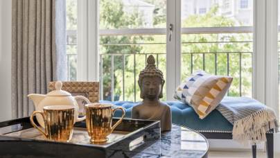 A living room with a marble coffee table featuring a tray with two gold mugs and a teapot, a Buddha statue, a bench with cushions, and large windows showing greenery outside.