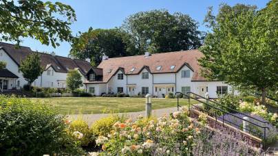 A large white house with a reddish-brown roof is surrounded by gardens, trees, and flowering plants under a clear blue sky.