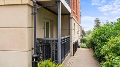 Ground-level apartment balcony with metal railing beside a paved walkway, surrounded by greenery and bordered by a brick and stone building.