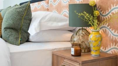 A bedside table with a green lamp, a yellow vase with flowers, and a candle next to a bed with white sheets and green pillows. Geometric headboard in background.
