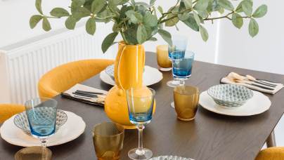 A dining table set with patterned plates, textured glasses, silverware, and a yellow vase with green foliage, surrounded by mustard-colored chairs.